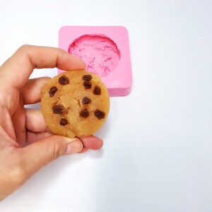 May include: A round, light brown cookie with dark brown chocolate chips is held in front of a pink silicone mold. The cookie is a soap or bath product, and the mold is in the shape of a cookie.