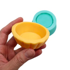 Könnte beinhalten: Eine gelbe Silikonform zum Backen von kleinen Kuchen oder Desserts. Die Form hat einen geriffelten Rand und einen glatten, runden Boden. Die Form wird neben einer türkisfarbenen Silikonform gezeigt.