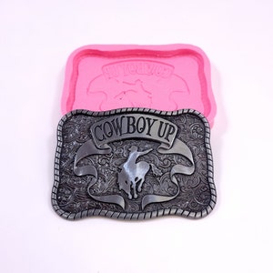 May include: A silver-toned metal belt buckle with an ornate design featuring a cowboy riding a bucking bronco. The buckle has the words "COWBOY UP" inscribed on a banner.