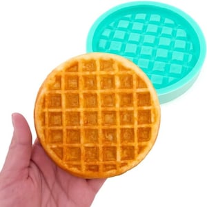 May include: A freshly made, golden-brown waffle with a square grid pattern, held in a hand. A teal silicone waffle mould is in the background. The waffle is a delicious treat, ready to be enjoyed.