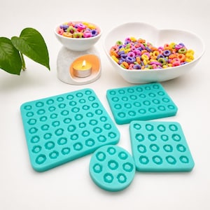 May include: Four turquoise silicone molds in square, rectangular, and circular shapes, each featuring multiple small ring-shaped cavities. These flexible crafting tools are arranged alongside a white ceramic wax warmer with a lit tea light candle.