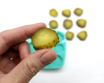 May include: A hand holding a single pickle slice, with a teal silicone mold in the background containing multiple pickle slices.
