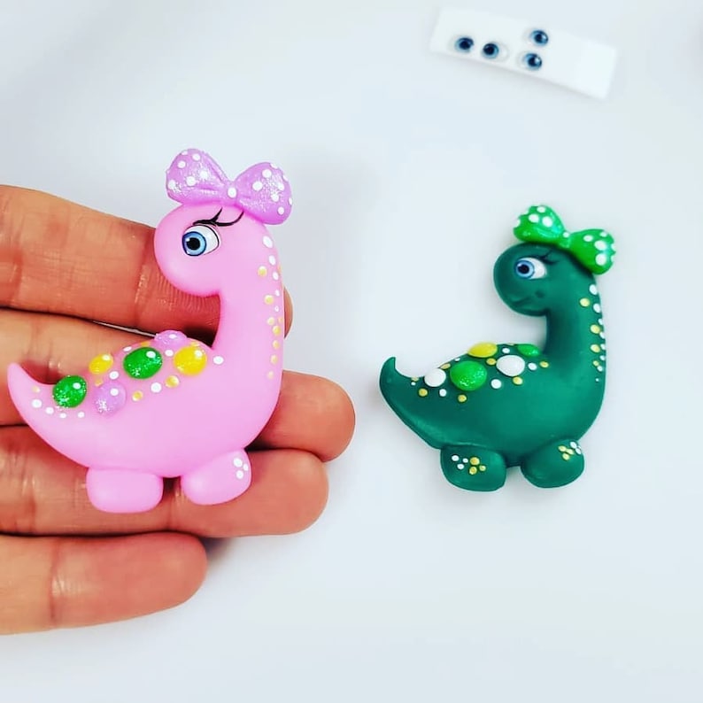May include: Two small dinosaur figurines, one pink and one green, with white polka dots and green and yellow accents. Both dinosaurs have a bow on their head and googly eyes.
