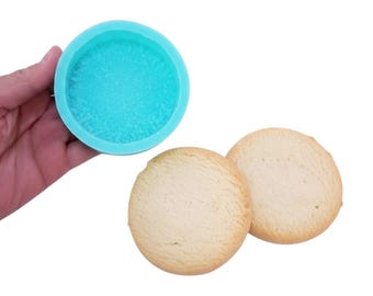 May include: A light blue silicone mold and two round, light-colored cookies are displayed against a white background. The mold is circular and has a textured interior. The cookies have a slightly rough surface.