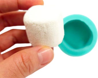 Può includere: Un marshmallow bianco cilindrico tenuto in mano, accanto a uno stampo in silicone color turchese. Il marshmallow ha un aspetto morbido e strutturato. Lo stampo è rotondo con un interno liscio.
