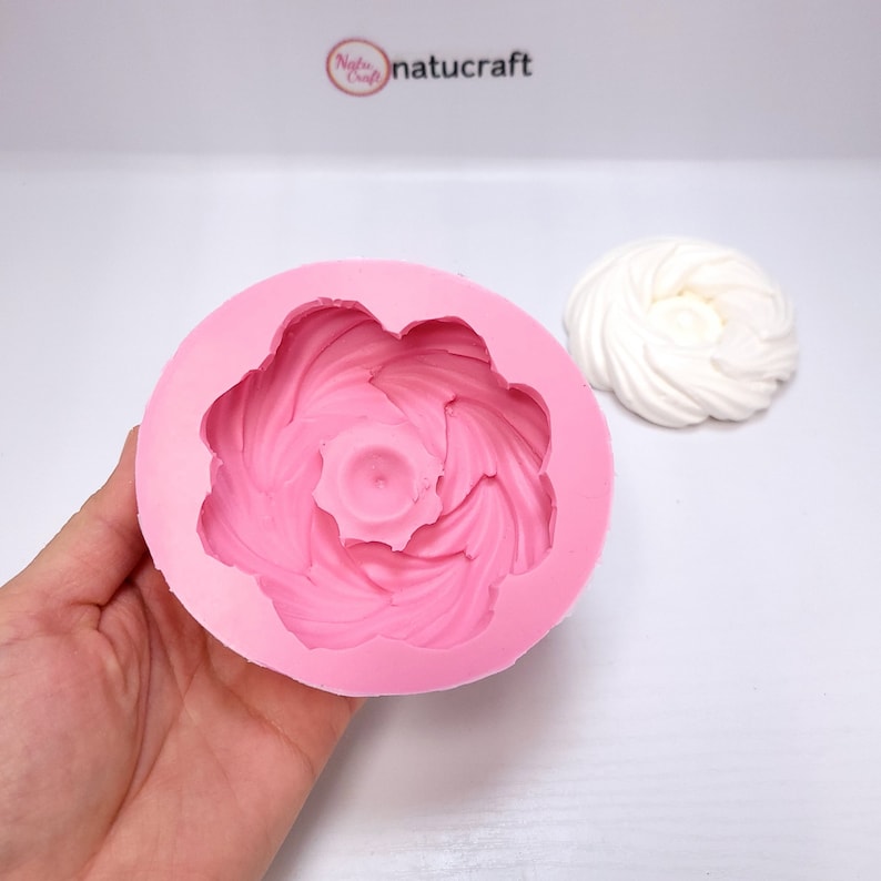 Op de afbeelding: Een roze siliconen mal in de vorm van een bloem, vastgehouden in een hand. De mal heeft een gedetailleerd, wervelend ontwerp. Een witte, bloemvormige lekkernij staat naast de mal. De merknaam "Natucraft" staat op de achtergrond.