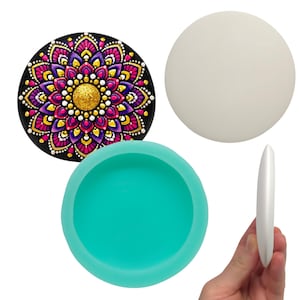 May include: A collection of craft supplies including a black, round, painted mandala with gold, pink, purple, and yellow details. Also included are a white, round object, a teal-colored silicone mold, and a white, elongated tool.