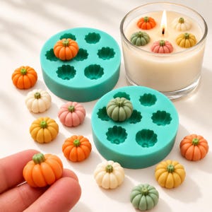 Op de afbeelding: Twee ronde turquoise siliconen mallen voor het gieten van kleine pompoenvormen, vergezeld door talloze miniatuur pompoenfiguurtjes in oranje, geel, wit, roze en saliegroen, plus een glazen kaars met diverse pompoenen op het kaarsvet.