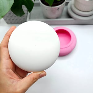 Könnte beinhalten: Eine weiße runde Form zum Herstellen eines kugelförmigen Objekts. Die Form ist offen und im Hintergrund ist eine rosa Silikonform zu sehen.