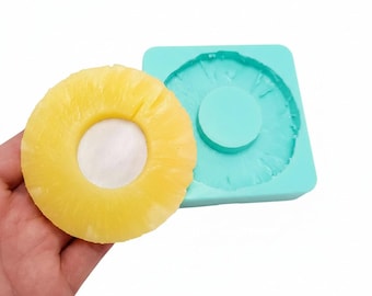 May include: A bright yellow pineapple slice next to a turquoise silicone mold. The pineapple slice has a white center and a textured, ring-shaped appearance. The mold is square with a circular indentation in the center.