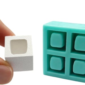 Op de afbeelding: Een klein, wit, vierkant object met een hol midden wordt door een vinger vastgehouden. Daarnaast een turquoise, vierkante mal met vier verzonken, afgeronde vierkante holtes. De objecten staan tegen een witte achtergrond.