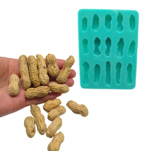 May include: A rectangular turquoise silicone mold featuring fifteen recessed cavities shaped like peanuts, positioned next to a collection of light brown, textured, whole peanuts in their shells, showcasing the mold's design and scale.