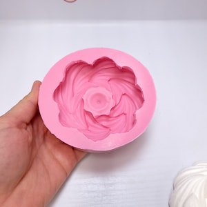 Op de afbeelding: Een roze siliconen mal in een cirkelvorm, met een gedetailleerd bloemenontwerp. De mal heeft een centraal, verhoogd element omgeven door wervelende bloembladachtige patronen. Het algehele ontwerp doet denken aan een roos of een andere bloem.
