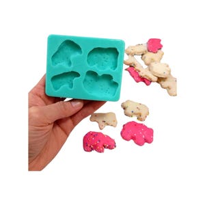 May include: A turquoise silicone mould with four bear-shaped cavities. Several bear-shaped sweets are scattered around, some with pink frosting and sprinkles, others with white frosting and sprinkles. The sweets appear to be homemade.
