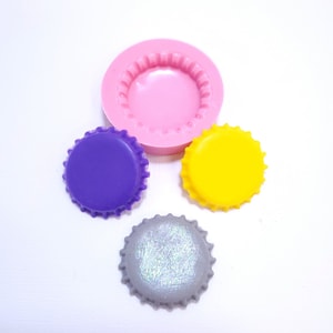 May include: Three bottle cap molds, one purple, one yellow, and one gray with glitter. The molds are made of silicone and are used to create bottle cap shaped decorations.