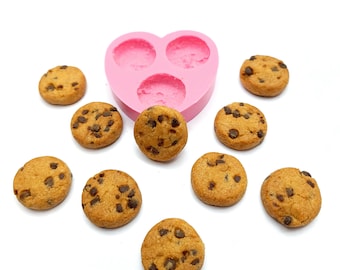 May include: A collection of miniature chocolate chip cookies scattered around a pink heart-shaped silicone mold. The cookies are golden brown with dark chocolate chips, and the mold has three circular cavities.