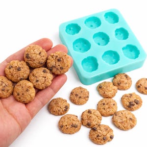 Pode incluir: Mini biscoitos com pepitas de chocolate, alguns na mão e outros espalhados numa superfície branca. Também está presente uma forma de silicone turquesa com nove cavidades circulares.