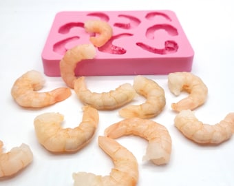 Può includere: Uno stampo in silicone rosa con 12 cavità a forma di gamberi. Ci sono 10 gamberi cotti su una superficie bianca accanto allo stampo.