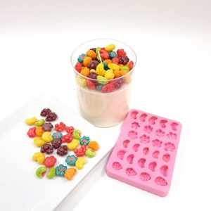 Pode incluir: Um frasco de vidro transparente cheio de velas com formato de cereais coloridos. Um molde de silicone rosa com várias formas de frutas e flores está ao lado do frasco. Um prato branco com velas com formato de cereais espalhadas está no fundo.
