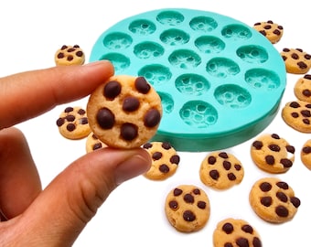May include: A close-up of a chocolate chip cookie held between fingers, with a teal silicone mould in the background. Several other cookies are scattered around the mould. The cookies have a light brown colour with dark brown chocolate chips.
