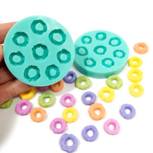 May include: Two teal silicone molds for making small donut-shaped treats. The molds are filled with small, colorful, donut-shaped cereal.
