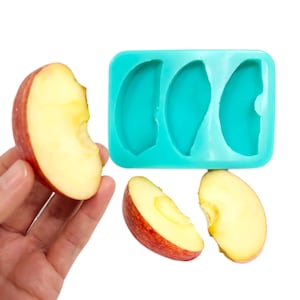 Peut inclure: Un moule en silicone turquoise avec trois formes en demi-cercle. Trois tranches de pomme rouge sont à côté du moule.