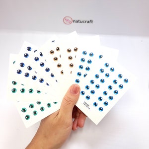 Puede incluir: Hojas de pegatinas de ojos en varios colores, incluyendo azul, verde y marrón. Cada hoja está llena de pequeños ojos de estilo caricatura. Las pegatinas se sostienen en una mano, con la marca "natucraft" visible en el fondo.