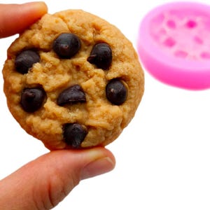 Puede incluir: Primer plano de una galleta con chispas de chocolate sostenida por una mano. La galleta es redonda, de color marrón dorado y con chispas de chocolate oscuro. Un molde de silicona rosa está en el fondo.