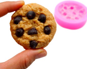 May include: A close-up of a chocolate chip cookie held by a hand. The cookie is round, with a golden-brown colour and dark chocolate chips. A pink silicone mould is in the background.