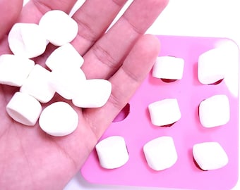 Può includere: Una mano che tiene diversi marshmallow bianchi accanto a uno stampo in silicone rosa con cavità a forma di marshmallow. I marshmallow sono morbidi e soffici e lo stampo ha nove cavità.