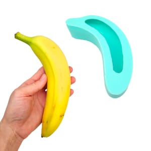 Könnte beinhalten: Eine reife gelbe Banane neben einer türkisfarbenen Silikonform in Bananenform. Die Banane hat einen grünen Stiel und ist leicht gebogen. Die Form ist einfarbig und hat einen hohlen Innenraum, der die Form der Banane nachbilden soll.