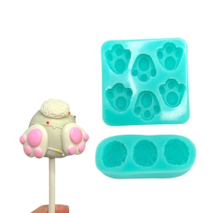 May include: A set of two turquoise silicone molds for making cake pops. One mold has six paw print shapes and the other has three round shapes. A cake pop with a white and gray bunny tail is shown.