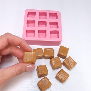 Peut inclure: Un moule en silicone rose avec 12 cavités carrées, et neuf caramels dans différentes positions sur une surface blanche.