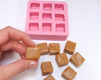 May include: A pink silicone mold with nine square cavities, next to several caramel candies. The candies are a light brown color and have a textured surface. One candy is being held by a hand. The image is on a white background.