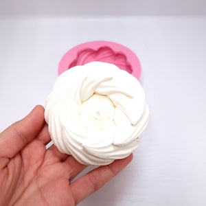 Op de afbeelding: Een wit, spiraalvormig dessert, mogelijk meringue, wordt in een hand gehouden, met een roze siliconen mal op de achtergrond. Ideaal om te bakken of te decoreren. Het dessert is een licht, luchtig gebak.