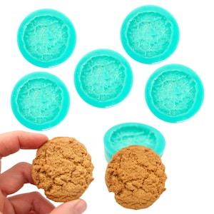May include: A close-up shot shows six turquoise silicone molds in a circular shape, with a textured interior. Two brown cookies are also visible. One cookie is held by a hand. The molds are designed for baking or crafting.