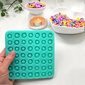 Könnte beinhalten: Eine quadratische, mintgrüne Silikonform mit einem Raster aus neunundvierzig kleinen, kreisförmigen Vertiefungen zur Herstellung von ringförmigen Miniaturartikeln. Die flexible Form hat abgerundete Ecken und eine matte Oberfläche.