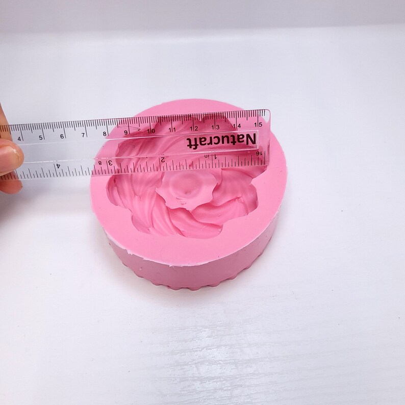 Op de afbeelding: Een roze siliconen mal in de vorm van een bloem, met een liniaal die een diameter van ongeveer 10 cm aangeeft. De mal heeft de tekst "Natucraft" erop.