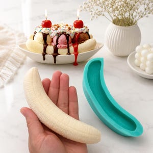 Puede incluir: Un molde de silicona flexible de color verde azulado con forma de plátano, acompañado de un vaciado realista de cera o jabón. El molde presenta un diseño curvo y texturizado, mientras que el vaciado muestra crestas detalladas que imitan un plátano.