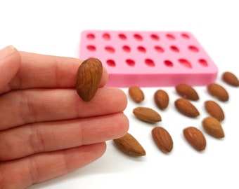May include: A pink silicone mold with 24 individual cavities shaped like almonds. A hand is holding a single almond, and several almonds are scattered on a white surface.