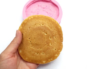 May include: A silicone mould for making a pancake with a circular design. The mould is pink and the pancake is brown and has a textured surface.