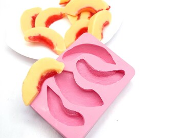 May include: Pink silicone mold with four peach slice shapes. A peach slice is sitting on top of the mold.