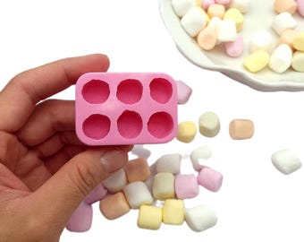 Può includere: Un piccolo stampo in silicone rosa con sei cavità circolari è tenuto in mano. Intorno, ci sono marshmallow colorati in tonalità di bianco, rosa, giallo e pesca. I marshmallow sono su una superficie bianca.