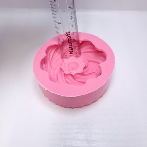 Op de afbeelding: Een roze siliconen bloemvormige mal met een centraal bloemontwerp. Een liniaal is over de mal geplaatst, met een diameter van ongeveer 5 cm. De mal is ontworpen voor knutselen of bakken, met een gedetailleerd bloemenpatroon.