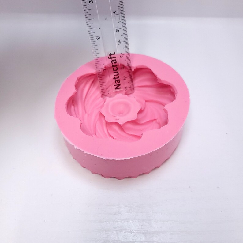 Op de afbeelding: Een roze siliconen bloemvormige mal met een centraal bloemontwerp. Een liniaal is over de mal geplaatst, met een diameter van ongeveer 5 cm. De mal is ontworpen voor knutselen of bakken, met een gedetailleerd bloemenpatroon.