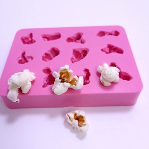 Pode incluir: Forma de silicone rosa com 10 cavidades em forma de pipoca. Existem quatro pedaços de pipoca fora da forma.
