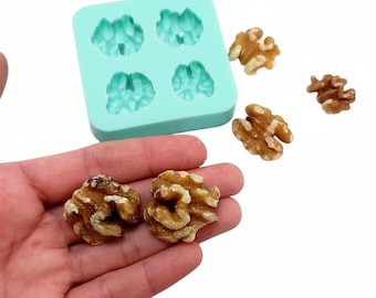 May include: A light blue silicone mold with four walnut-shaped cavities, alongside several shelled walnuts. Two walnuts are held in a hand. The walnuts are light brown and have a textured surface. The mold is square and has a smooth surface.