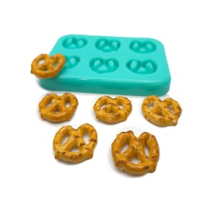 Puede incluir: Un molde de silicona turquesa con seis cavidades en forma de corazón para hacer pretzels en miniatura. Seis pretzels en miniatura están esparcidos alrededor del molde.