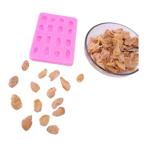 May include: A pink silicone mould with oval-shaped cavities, scattered candied ginger pieces, and a glass bowl filled with candied ginger. The ginger pieces are light brown and have a crystallized coating. The mould is designed for creating shaped treats.