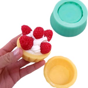 May include: A handmade soap resembling a raspberry tart, featuring red raspberries and white cream on a yellow pastry base. A teal silicone mould and a yellow mould are also visible.